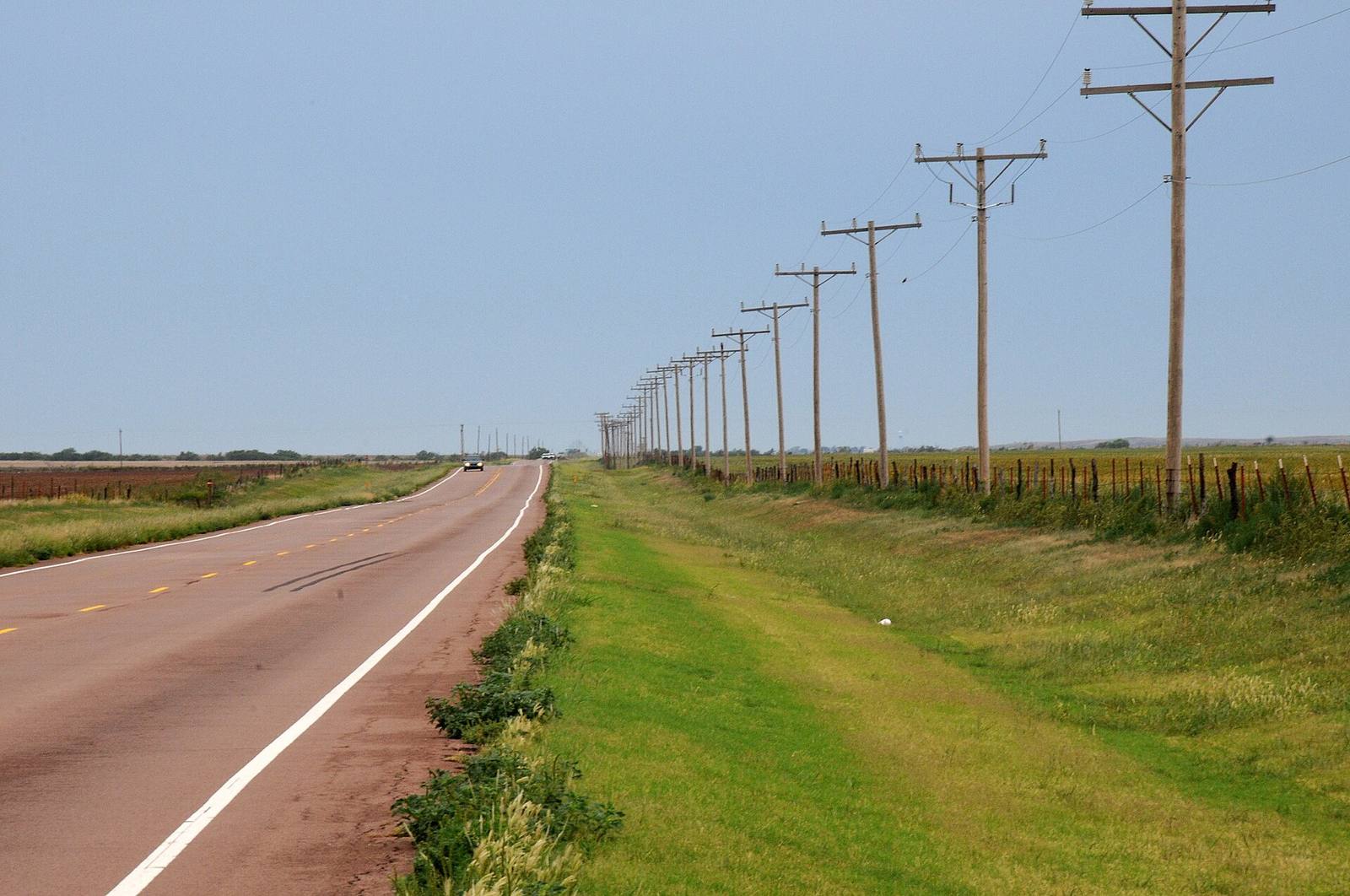 A line of wooden distribution poles running beside an empty rural highway across open farmland