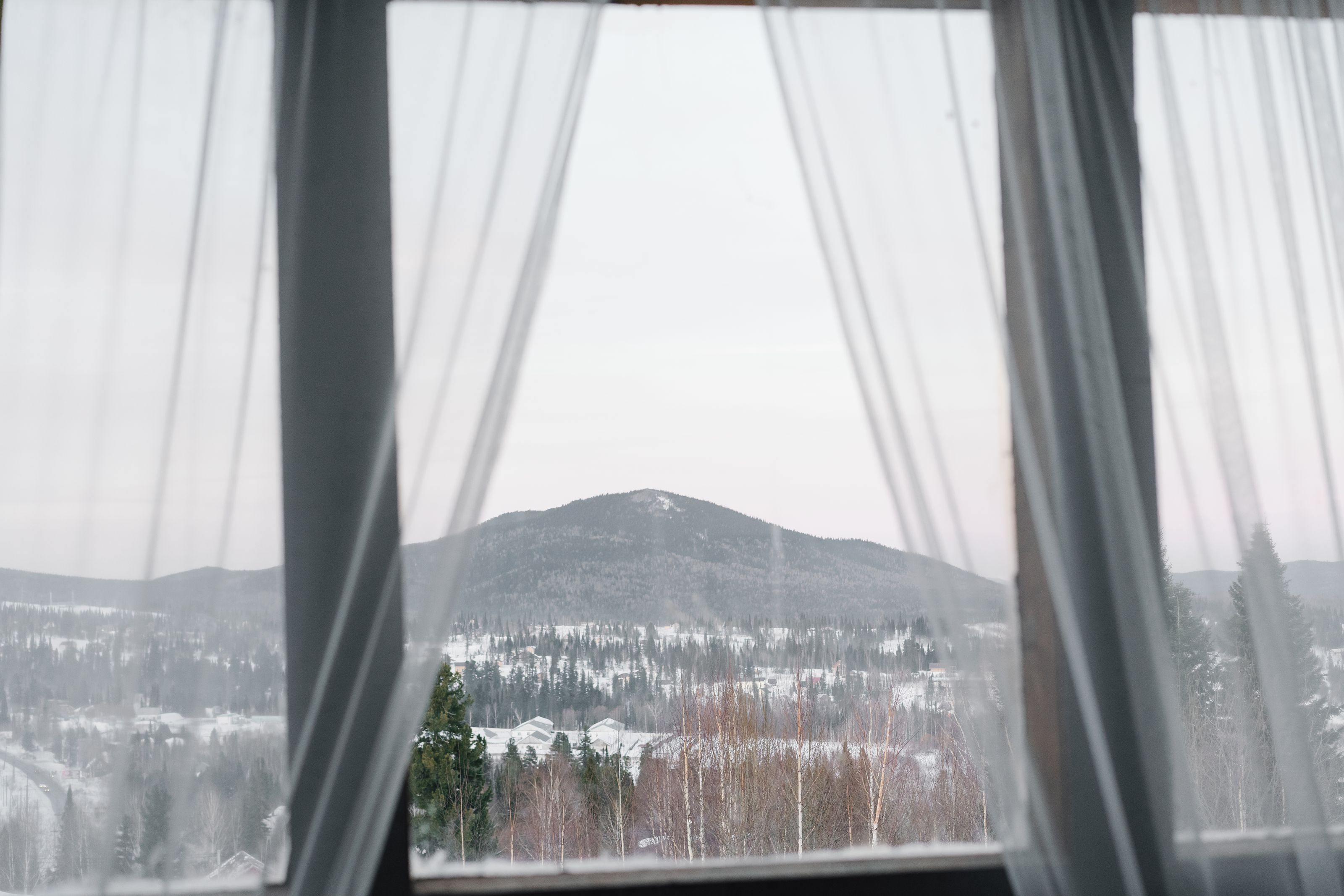 Sheer curtains at a cabin window opening onto snow-covered hills and forest