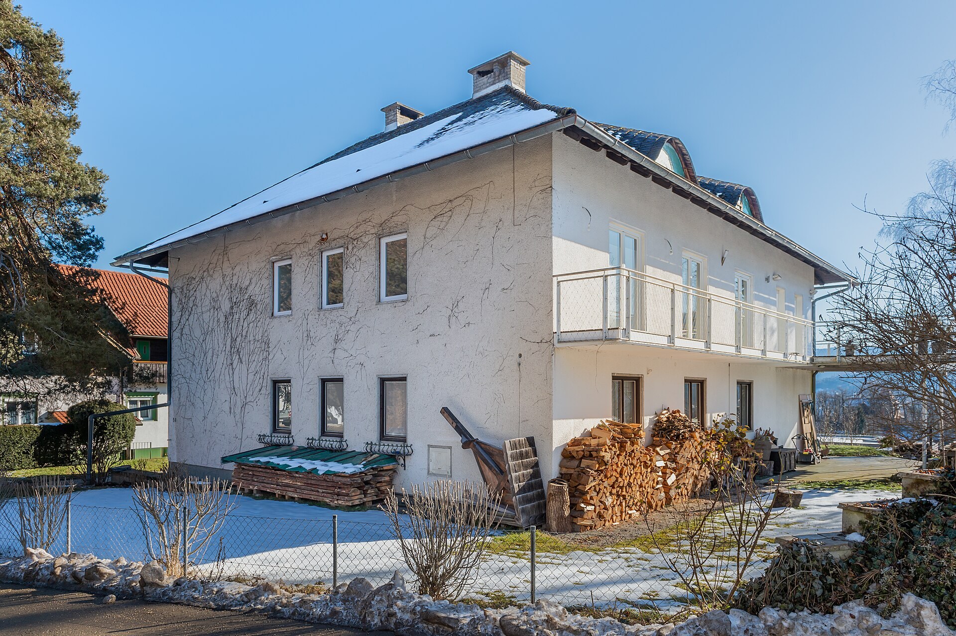 Snow on the roof of a rendered house with a tall stack of split firewood piled against its wall in winter sunshine