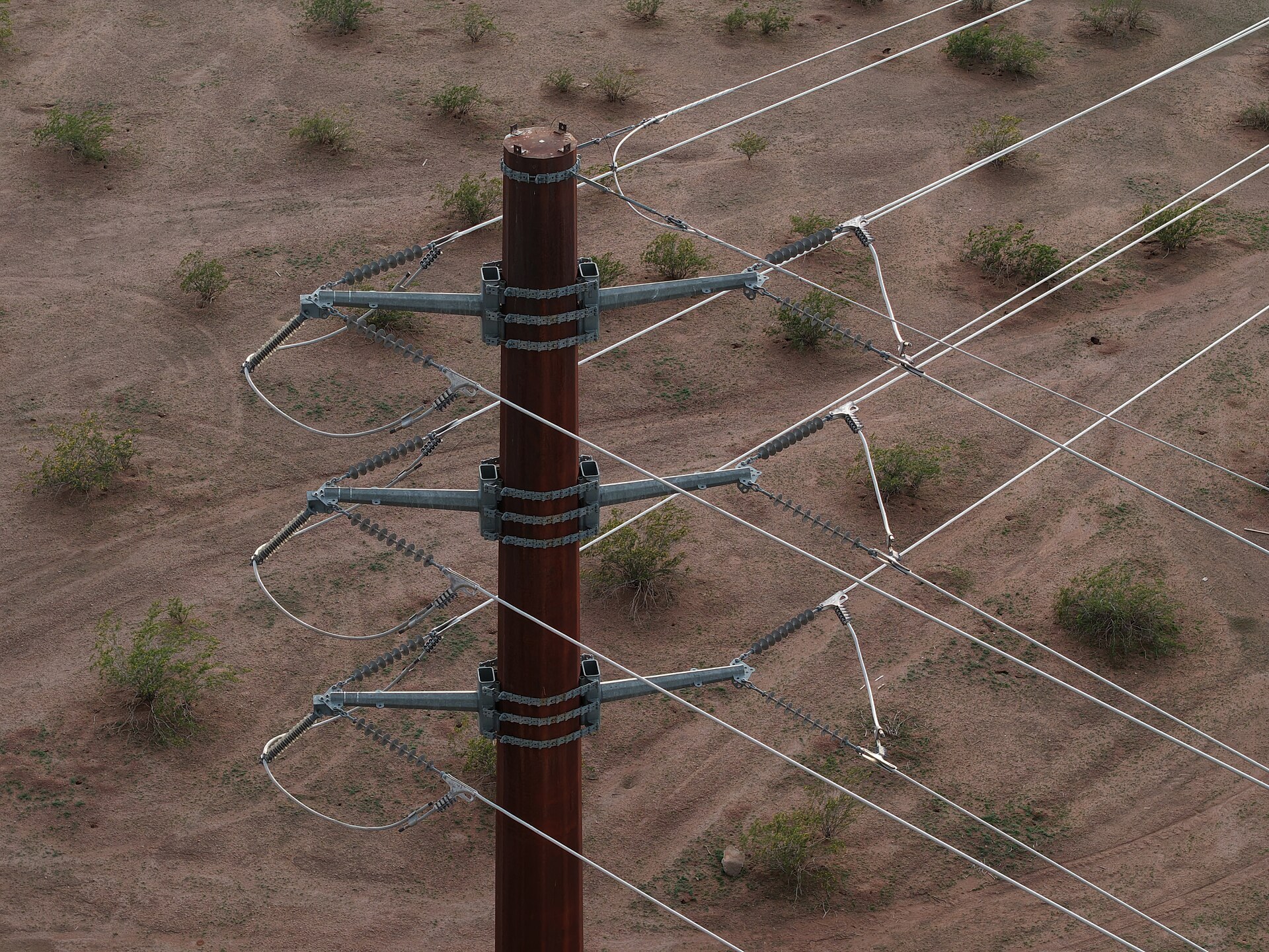 Aerial view of a steel transmission monopole carrying three phases of bare conductor across open desert ground