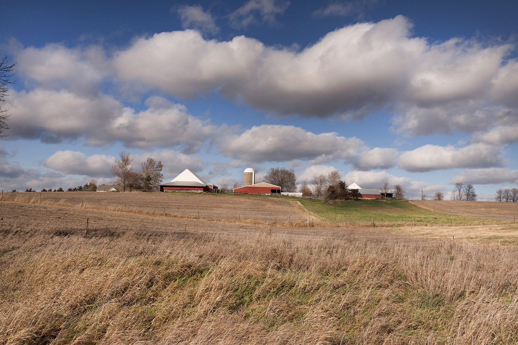 Open farmland running to a low ridge, with a farmstead and grain silo on the horizon under a broken sky