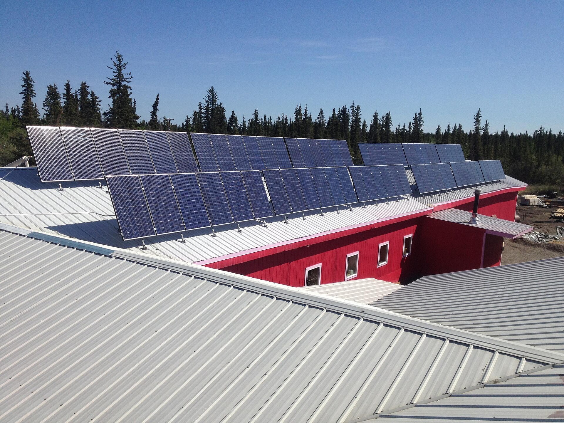 Solar panels tilted steeply on the metal roof of a red building at the edge of a spruce forest under a clear sky