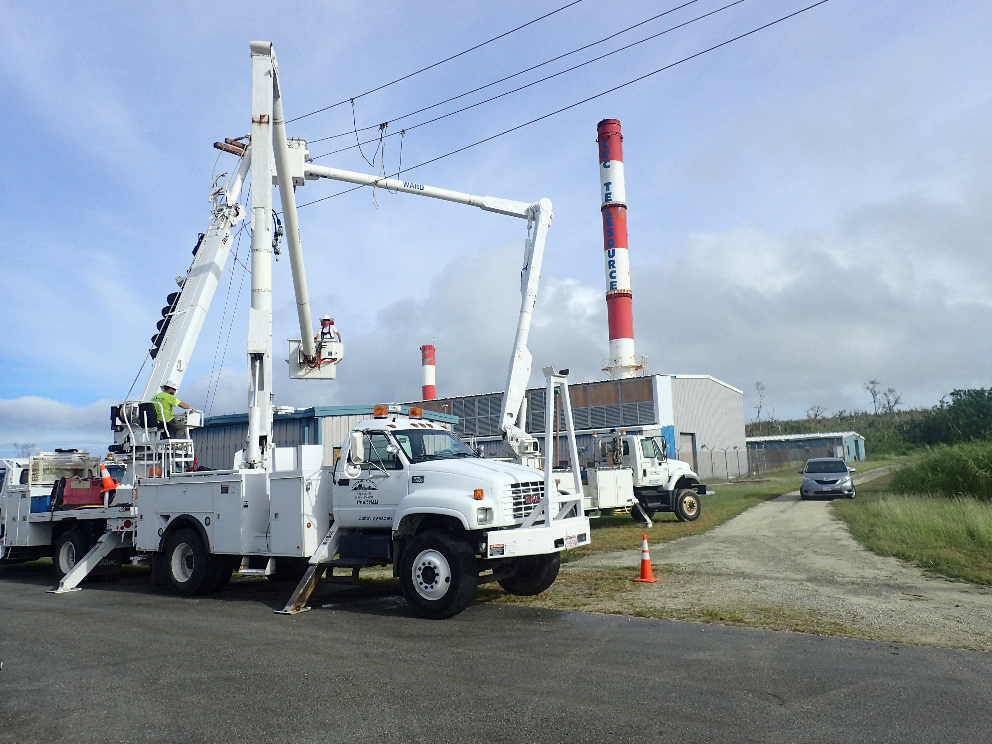 Utility bucket trucks and a line crew rebuilding storm-damaged distribution lines beside a power plant