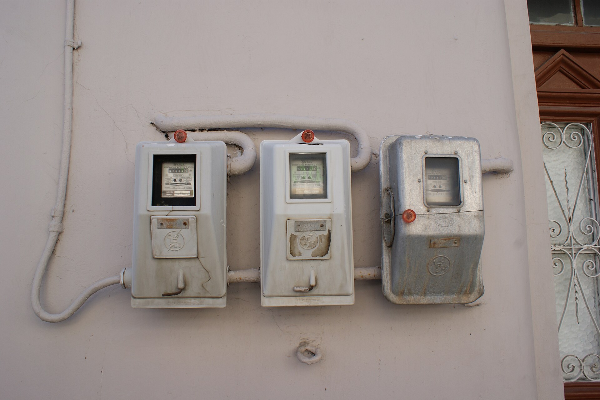 Three old electricity meters mounted side by side on a whitewashed house wall with surface-run conduit