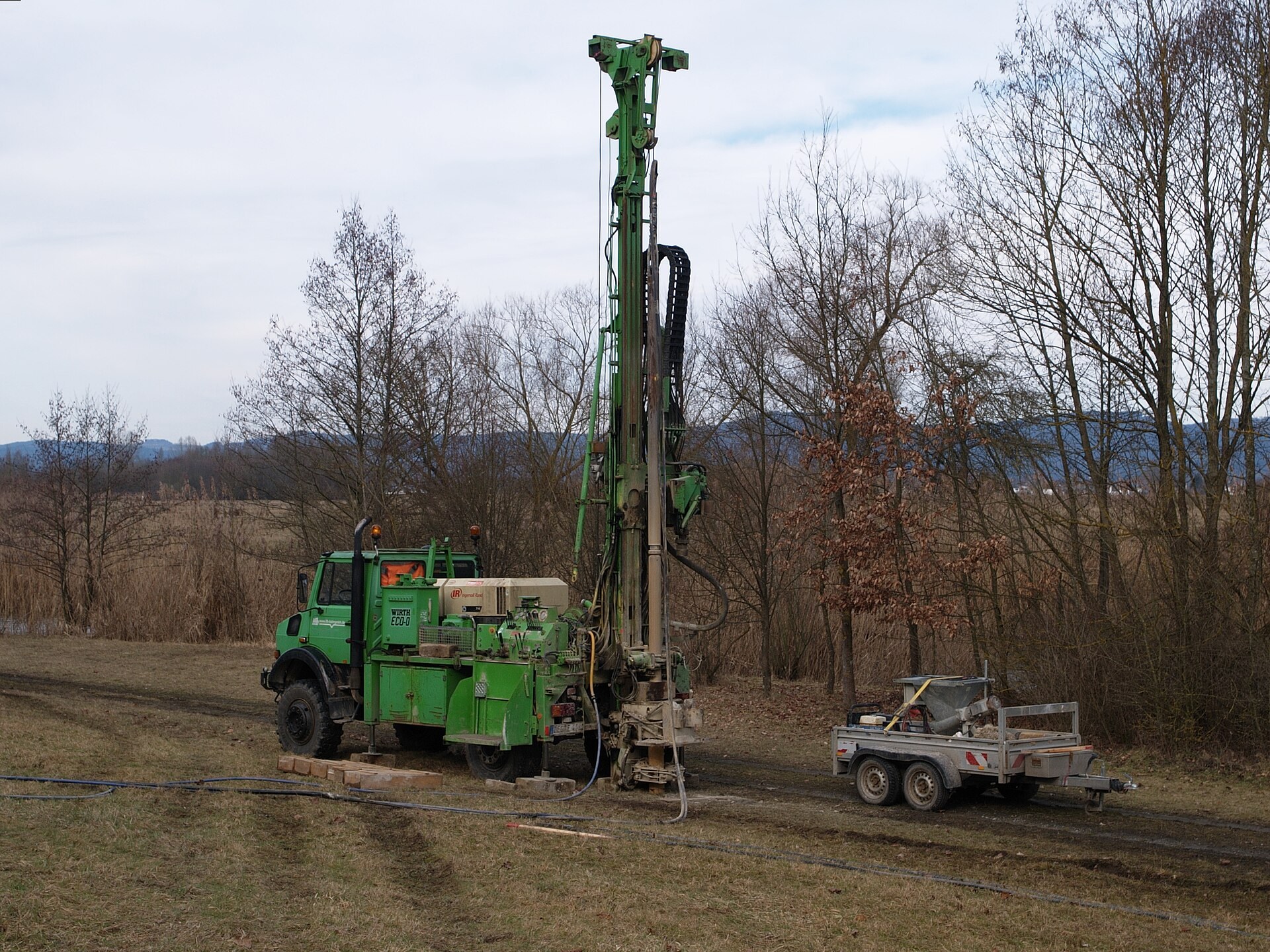 Truck-mounted drilling rig with its mast raised, drilling a water borehole in a bare field beside a line of leafless trees
